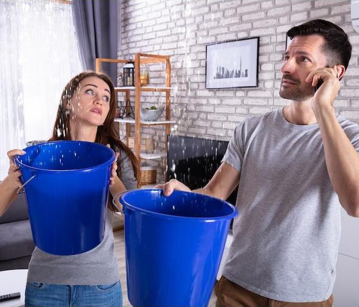 Couple Using Bucket For Collecting Water Leakage From Ceiling And Talking On Cellphone
